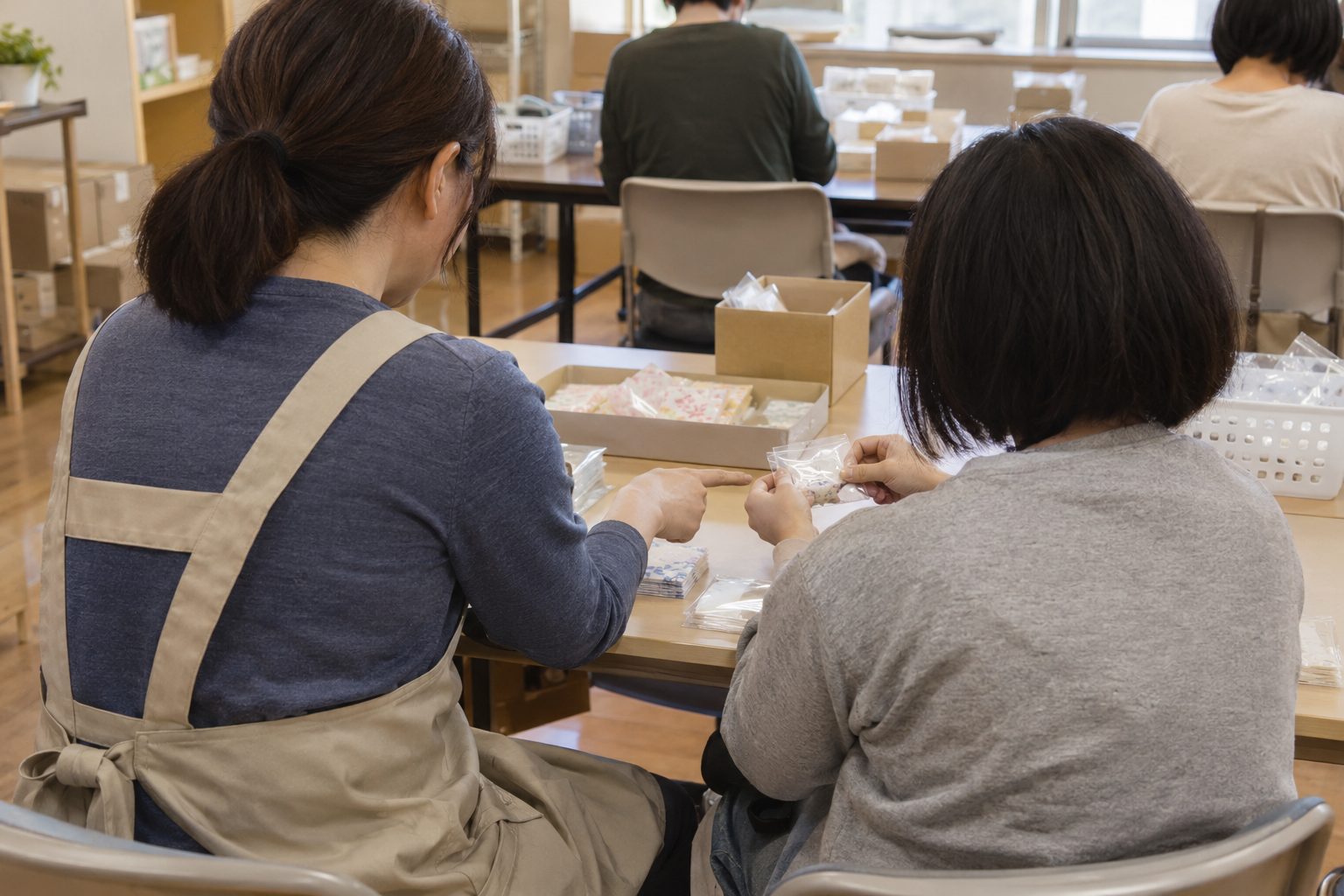 職員が利用者の隣に座り、袋入れの作業を指でさしながら一緒に進めている後ろ姿