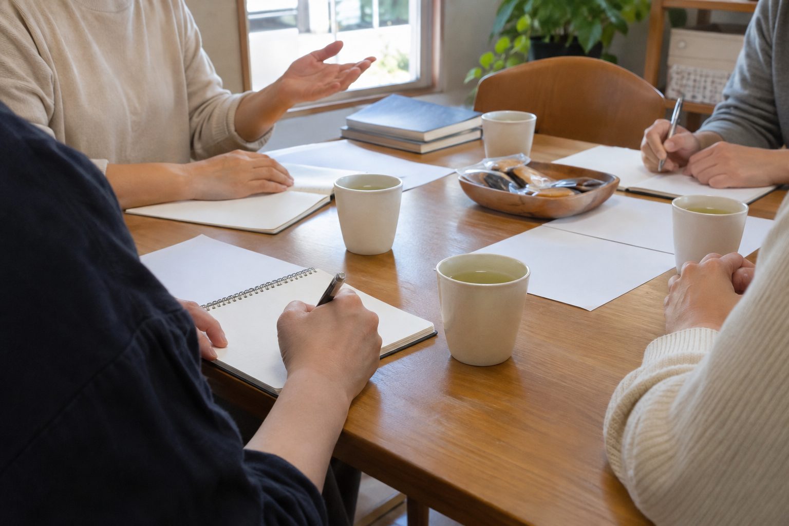 話し合いの机を囲む人たちの手元。手のひらを上に向けて話す人と、ノートにペンで書き留める人。白紙のノートと紙、緑茶の湯のみ、お菓子の皿